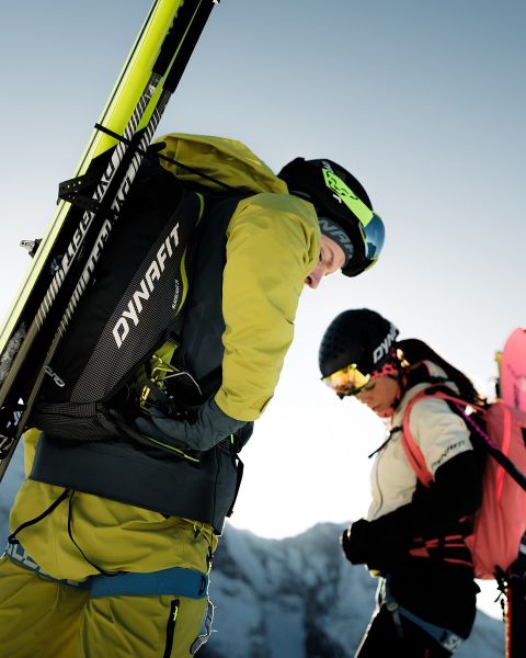 Dos personas corren por un sendero de montaña con una ciudad y un río al fondo. Uno lleva una camiseta negra y pantalones cortos, el otro una camiseta de tirantes azul y pantalones cortos. El logo de Dynafit está en la parte superior de la imagen.