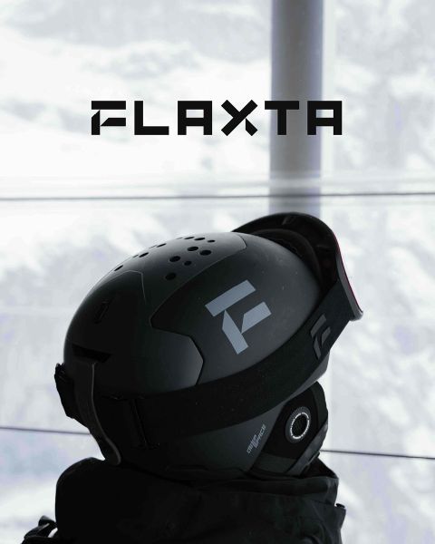 Dos esquiadores con cascos y equipo están de pie en una plataforma de metal cubierta de nieve con vistas a un paisaje montañoso nevado. Sostienen esquíes en sus manos y están uno frente al otro. Encima de ellos, la palabra "FLAXTA" destaca en letras grandes.