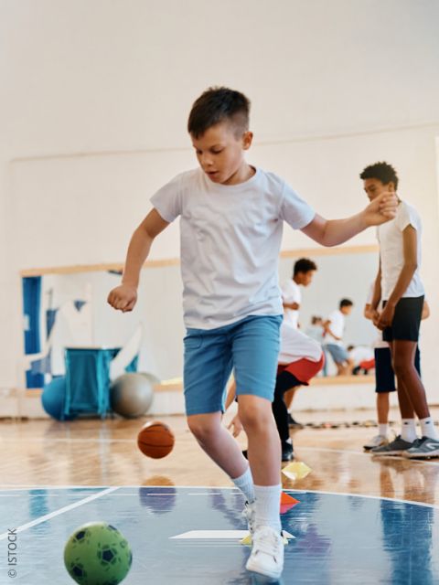 Un niño con una camiseta blanca y pantalones cortos azules se equilibra sobre un pie mientras juega en un gimnasio. Al fondo se ven otros niños y balones deportivos.