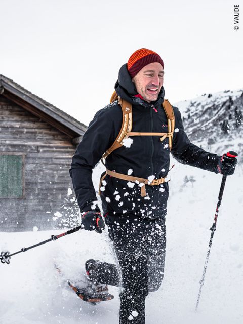 Una persona con ropa de invierno y un gorro rojo camina con raquetas de nieve a través de la nieve profunda cerca de una cabaña de madera, sosteniendo bastones de trekking en la mano y con la nieve arremolinándose a su alrededor.