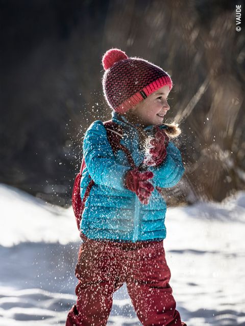 Un niño pequeño con ropa de invierno lanza nieve, sonríe y juega afuera en un día soleado y nevado. El niño lleva una chaqueta azul, pantalones rojos y un gorro de lana rojo con un pompón.
