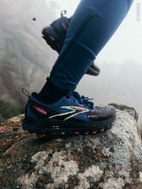 Una persona con zapatillas de trail running azules y leggings azules entra en una zona rocosa al aire libre, con niebla y rocas al fondo.