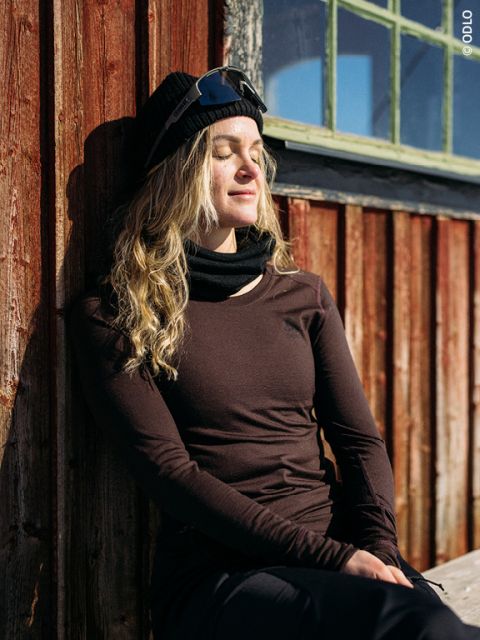 Una persona con cabello largo y rubio, gorra negra, gafas de sol y ropa oscura está sentada con los ojos cerrados junto a una pared de madera a la luz del sol, pareciendo relajada y satisfecha.