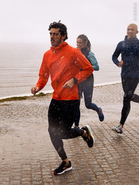 Tres personas trotan bajo la lluvia sobre un camino adoquinado mojado junto al agua. La persona del frente lleva una chaqueta roja, mientras que las dos personas de atrás visten ropa más oscura. La lluvia cae visiblemente a su alrededor.