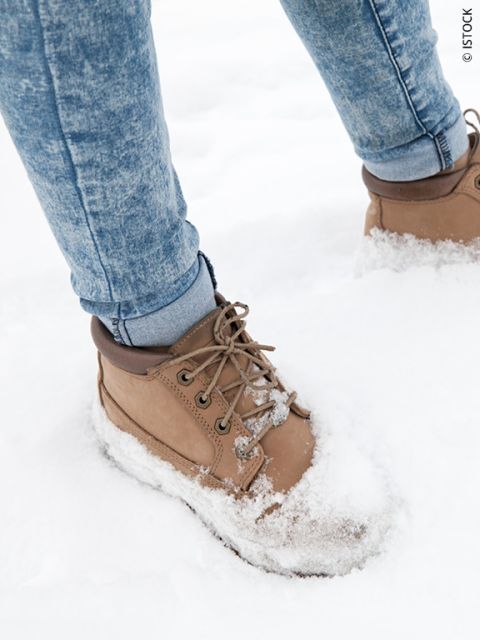 Una persona con botas de color marrón claro y jeans azules está de pie en la nieve blanca y fresca, con la nieve cubriendo las botas y parte de los jeans.