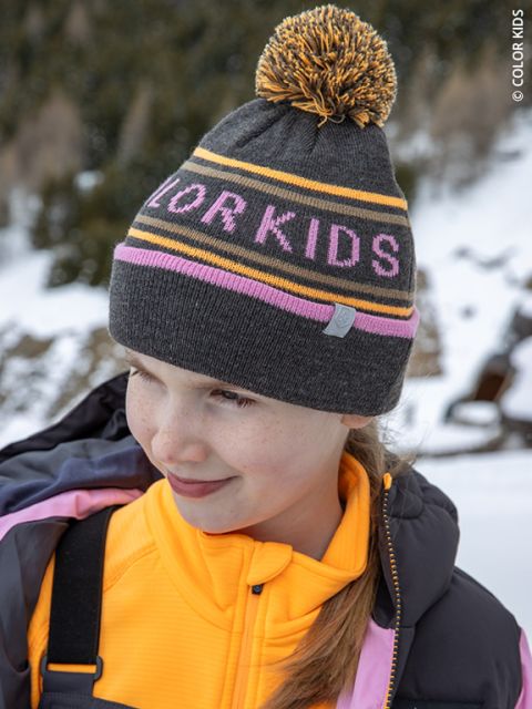 Un niño sonriente con un gorro gris que dice "COLOR KIDS" lleva una chaqueta amarilla y un abrigo morado, y está de pie al aire libre en un paisaje montañoso nevado.