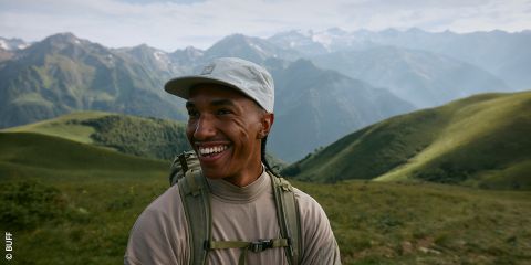 Una persona sonriente, con un gorro ligero y una mochila, está de pie al aire libre en un paisaje montañoso verde con picos distantes y brumosos bajo un cielo parcialmente nublado.