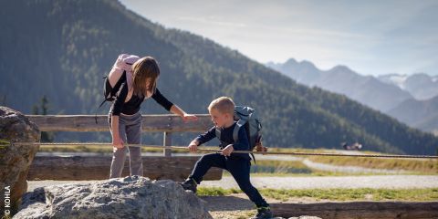 Una mujer ayuda a un niño a escalar rocas en una ruta de senderismo con una cuerda; ambos llevan mochilas, con montañas y árboles al fondo bajo un cielo despejado.