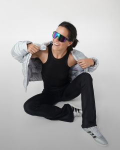 Una mujer está sentada en el suelo y se pone una sudadera gris por la cabeza sonriendo. Lleva una camiseta negra sin mangas, pantalones de chándal grises, calcetines blancos y zapatillas deportivas grises sobre un fondo liso de color gris claro.