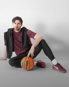 Un hombre con cabello castaño y barba está sentado en el suelo frente a un bloque blanco. Lleva una chaqueta gris claro, una camisa negra, pantalones de chándal oscuros, calcetines blancos y zapatillas grises, y sostiene una botella de agua en la mano.