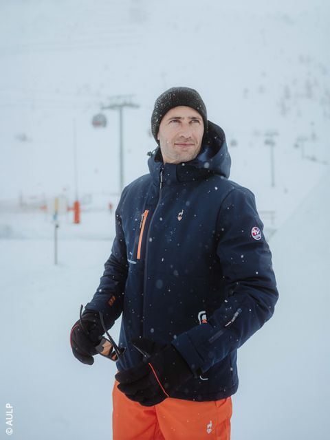 Un hombre con una chaqueta de invierno azul marino, pantalones naranjas, un gorro y guantes negros está de pie en un entorno nevado al aire libre sosteniendo unas gafas de sol. En el fondo se ven telesillas y pistas cubiertas de nieve.