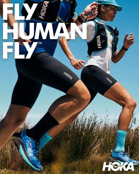 Dos corredores, un hombre y una mujer, corren por un sendero rocoso de montaña, rodeados de exuberante vegetación y altos árboles. Ambos llevan ropa de correr y mochilas de hidratación. En la parte superior se muestra claramente la palabra "HOKA". En el fondo se alzan montañas nevadas.