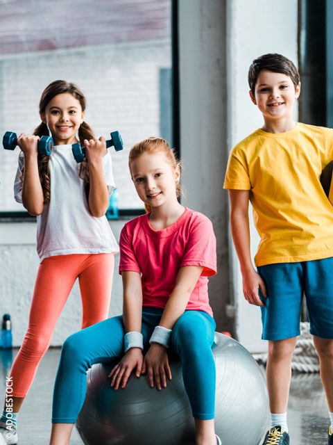 Tres niños sonrientes en ropa deportiva posan en un gimnasio. Una niña sostiene pesas, otra está sentada en una pelota de gimnasia y un niño está de pie junto a ellas. Parecen felices y listos para la actividad física.