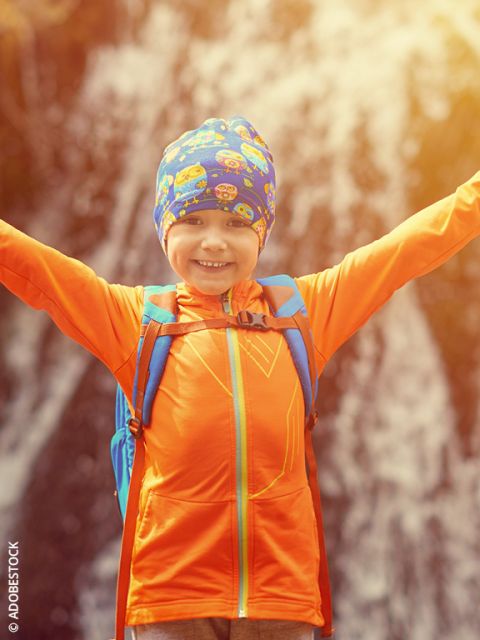 Niño sonriente con chaqueta naranja y mochila azul levanta los brazos frente a una cascada borrosa, y lleva un sombrero colorido con caras de dibujos animados. La luz del sol brilla en el fondo.