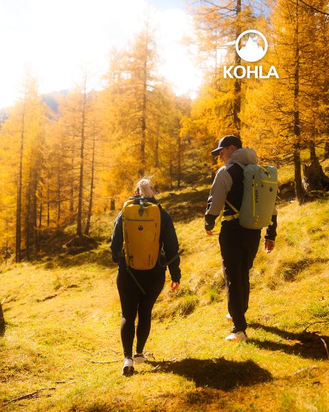 Dos excursionistas con mochilas caminan por un bosque soleado con árboles dorados de otoño. La luz del sol se filtra a través de las ramas, proyectando una luz cálida. El logo de Kohla se puede ver en la esquina superior derecha.