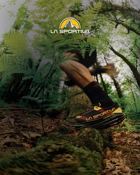 Cuatro personas con equipo de exterior y colchonetas de búlder a la espalda caminan por un sendero de montaña cubierto de hierba. Encima de ellos se ve el logotipo de La Sportiva en el cielo. Al fondo se ven escarpadas cumbres montañosas.