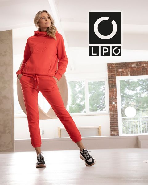 Un hombre con una camiseta azul y pantalones oscuros hace un ejercicio de cuerda de batalla en un gimnasio de estilo industrial con dos grandes ventanas de arco al fondo. La luz entra y las letras "LPO" aparecen en la esquina superior derecha.