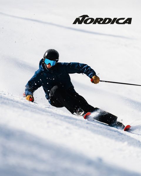 Dos esquiadores con equipo de protección, uno con ropa azul y el otro con ropa oscura, descienden una colina nevada. El logotipo de "Nordica" se ve en la parte superior. Ambos están en ángulo, demostrando velocidad y agilidad en un día soleado.