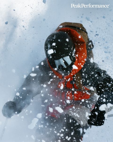 Una persona salta entre grandes rocas en una región montañosa bajo un cielo azul brillante. Lleva ropa deportiva, incluyendo una chaqueta y pantalones cortos. En la parte superior se puede ver la inscripción "Peak Performance".