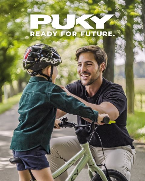 Una joven con casco azul y camiseta a rayas monta en bicicleta celeste en un día soleado. Sonriendo, pasa junto a coloridos grafitis en un entorno urbano. El logo de PUKY y el eslogan "Preparados para la vida" se ven en la parte superior izquierda.