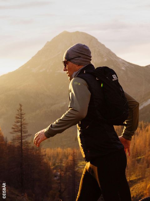 Una mujer con una chaqueta y guantes rosa grisáceos corre en un paisaje montañoso nevado, llevando dos botellas de agua en los bolsillos delanteros de su chaleco para correr.