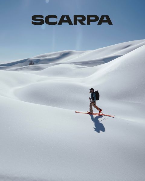 Una mujer y un hombre están sentados al aire libre sobre grandes rocas. Llevan ropa casual y gafas de sol. Están rodeados de vegetación y rocas, el cielo está ligeramente nublado. En la parte superior de la imagen se puede ver la palabra "SCARPA".