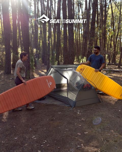 Una persona yace sobre una esterilla de camping, envuelta en un saco de dormir amarillo, y observa un amanecer sobre un acantilado costero. Equipo de camping está a su lado, y las olas del mar rompen contra las rocas. El logo "SEATOSUMMIT" se encuentra en la esquina inferior derecha.