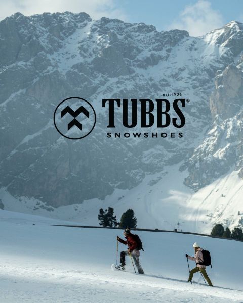 Dos personas caminando cuesta arriba por una ladera nevada con bastones de esquí, con altas montañas escarpadas y cubiertas de nieve al fondo. El logo de Tubbs Snowshoes aparece sobre la imagen.