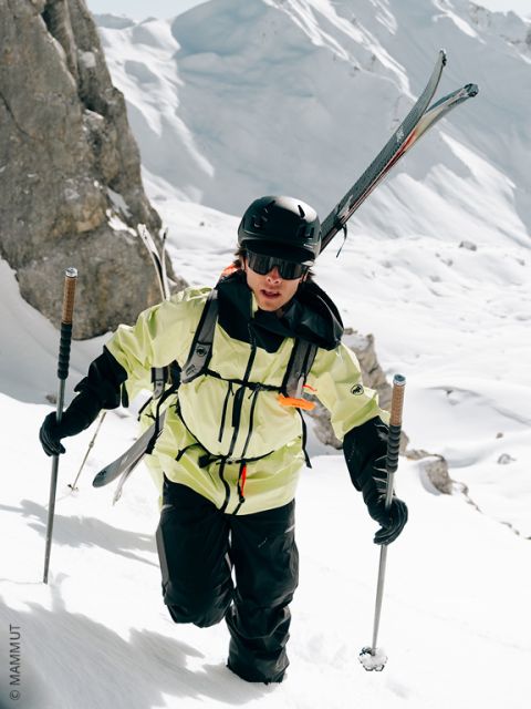 Un esquiador con casco, gafas y chaqueta amarilla sube por una ladera nevada, llevando los esquíes a la espalda y utilizando los bastones para apoyarse. Acantilados rocosos y nieve profunda rodean la escena.