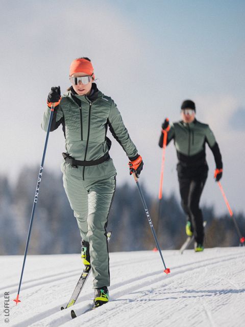 Dos personas vestidas de verde y negro con detalles naranjas practicando esquí de fondo en un paisaje nevado. La persona de delante lleva gafas de sol y una capucha; se ven árboles y un cielo azul al fondo.