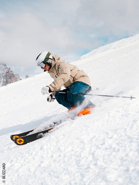Un esquiador con una chaqueta beige, pantalones azules y un casco blanco desciende por una pista nevada, haciendo un giro brusco y levantando nieve. En el fondo se ven montañas.