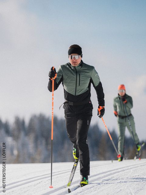 Dos esquiadores de fondo en una pista nevada, con equipo de deportes de invierno y gafas de sol, con árboles y cielo azul de fondo.