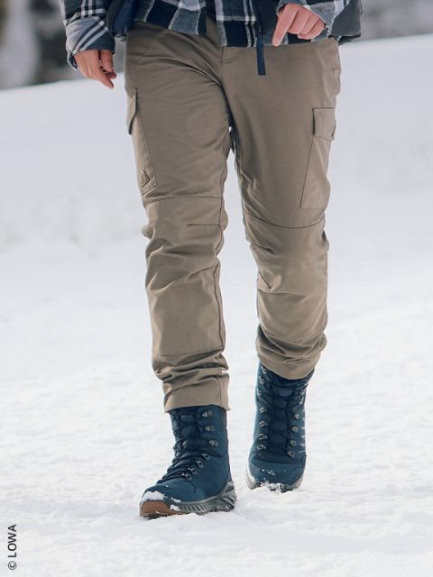 Persona con pantalones color caqui y botas de invierno azul marino caminando sobre la nieve, donde solo se ven sus piernas y parte de su torso.