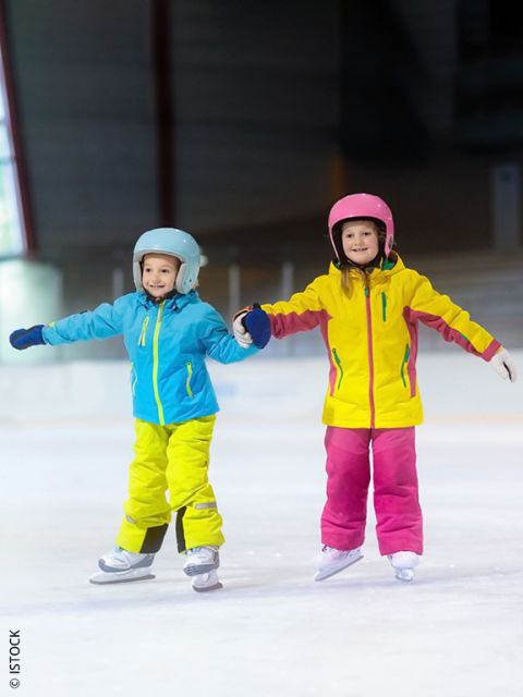 Dos niños pequeños con chaquetas y cascos coloridos patinan juntos, tomados de la mano y sonriendo en una pista de hielo.