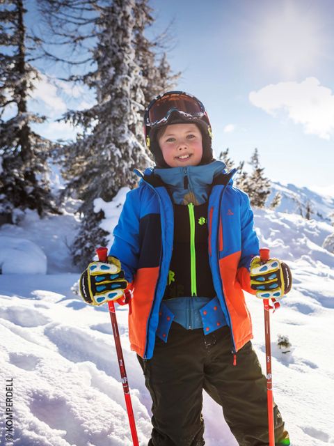 Niño sonriente con equipo de esquí y bastones de pie en un paisaje montañoso nevado con árboles y luz solar brillante al fondo.