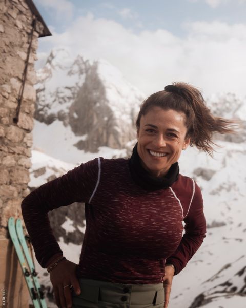 Una mujer sonriente con una blusa marrón castaño está de pie al aire libre cerca de un edificio de piedra con montañas nevadas y esquíes al fondo.
