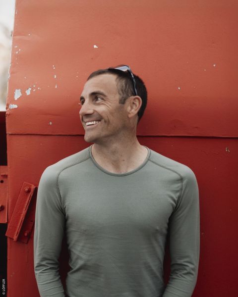 Un hombre sonriente con una camisa verde está de pie frente a una gran estructura metálica roja y mira hacia un lado con unas gafas de sol en la cabeza.