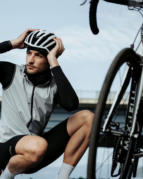 Un ciclista con casco y ropa de ciclismo se arrodilla junto a una bicicleta y se ajusta el casco, con un puente y el cielo de fondo.