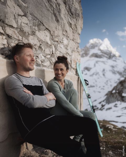 Dos personas con ropa de invierno están sentadas en la parte trasera de un vehículo estacionado en una calle nevada. Ambos están sonriendo. La persona de la izquierda lleva un gorro negro y una sudadera, mientras que la persona de la derecha lleva un jersey estampado, un casco y gafas protectoras. Texto en la imagen.