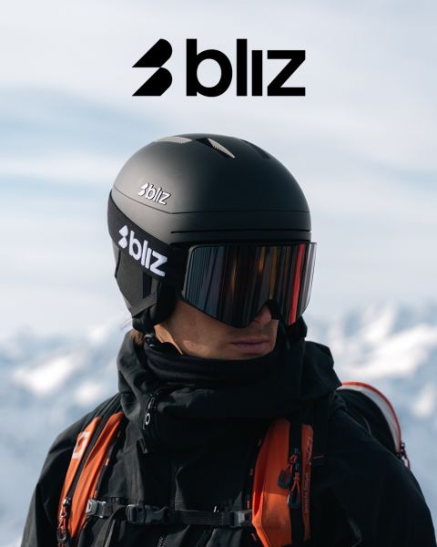 Una persona con un casco de esquí negro y gafas de esquí espejadas, ambos con la inscripción "Bliz", está de pie al aire libre con montañas nevadas al fondo. El logotipo de Bliz está colocado de forma destacada en la parte superior de la imagen.