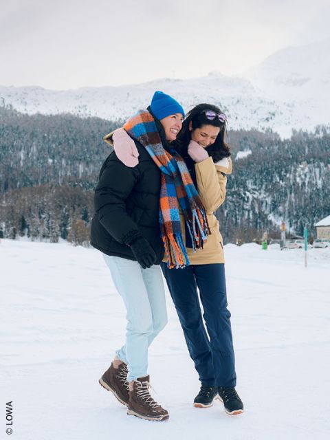 Dos mujeres con ropa de invierno ríen y caminan juntas por un paisaje nevado con montañas y árboles al fondo.