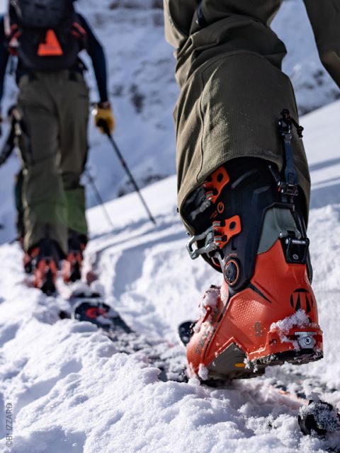 Primer plano de dos personas caminando cuesta arriba en la nieve con botas de esquí y bastones naranjas, con montañas nevadas al fondo.