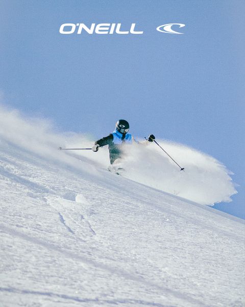 Una mujer con cabello largo está de pie en un paisaje desértico bajo un cielo azul con nubes dispersas. Lleva una camiseta sin mangas negra de O'Neill y se inclina ligeramente hacia adelante. En el lado derecho de la imagen se puede ver el logo de O'Neill.
