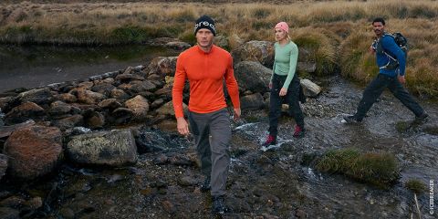 Tres personas con equipo para actividades al aire libre cruzan un arroyo rocoso en una zona montañosa cubierta de hierba, con una persona que va delante y las otras detrás.