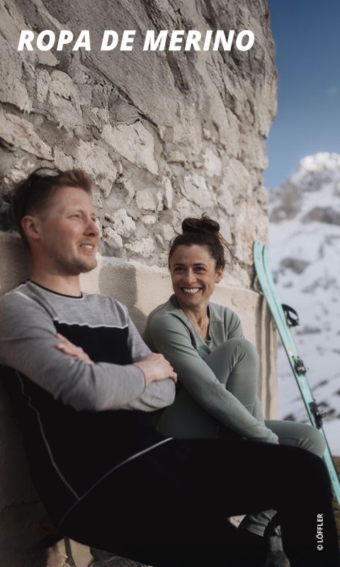 Un hombre sonriente y una mujer sonriente con ropa de merino sentados en una pared de piedra, cerca hay esquíes; al fondo se ven montañas nevadas.
