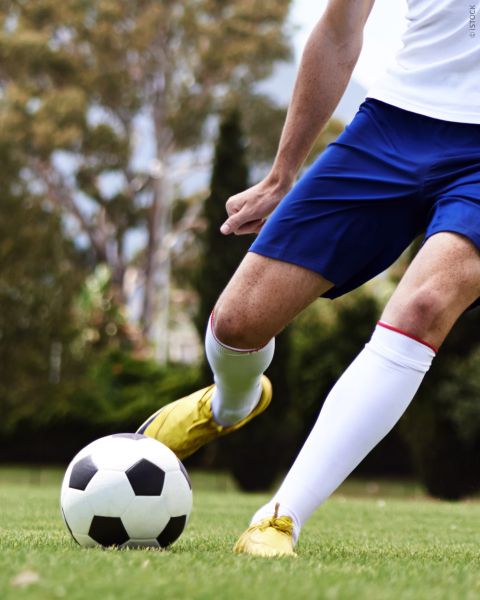 Futbolista en pantalones cortos azules y zapatos amarillos pateando un balón de fútbol blanco y negro en un prado, con árboles al fondo.