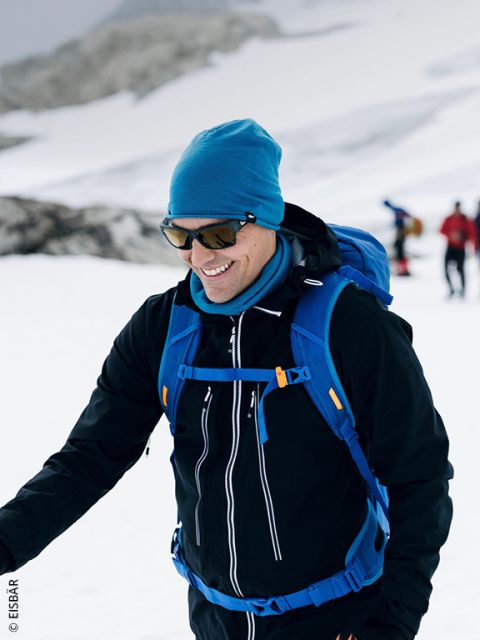 Una persona sonriente con ropa de invierno camina en un terreno nevado, lleva gafas de sol, un gorro azul y una mochila azul, mientras que en el fondo se ven otras personas.