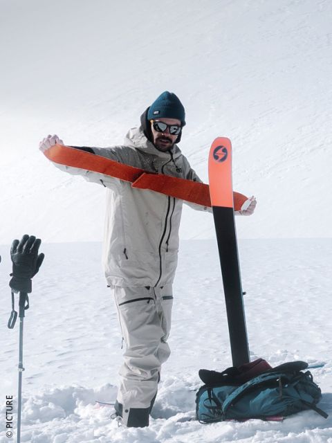 Una persona con equipo de invierno coloca una piel de foca naranja sobre un esquí en una montaña nevada, mientras lleva bastones de esquí y una mochila.