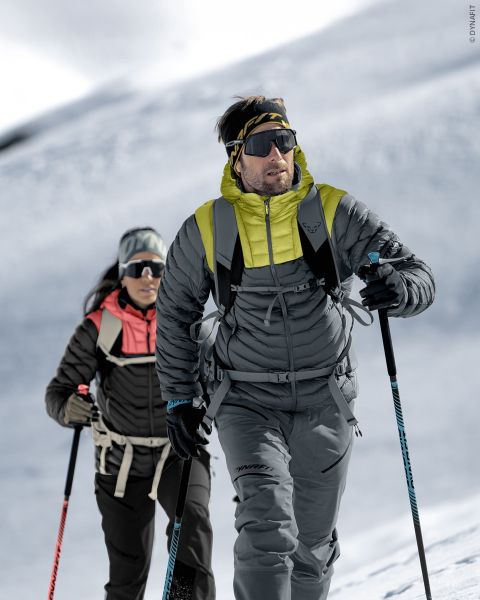 Dos personas con equipo de invierno caminan con bastones de esquí, gafas de sol y mochilas por una ladera nevada bajo un cielo brillante y nublado.