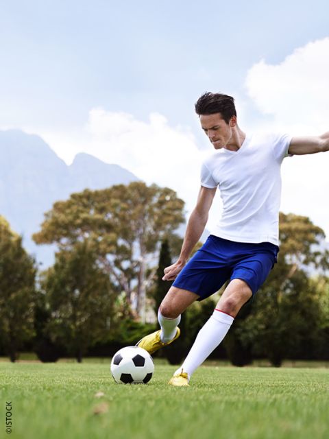 Un futbolista con una camisa blanca y pantalones cortos azules patea una pelota de fútbol en un césped con árboles y montañas al fondo.